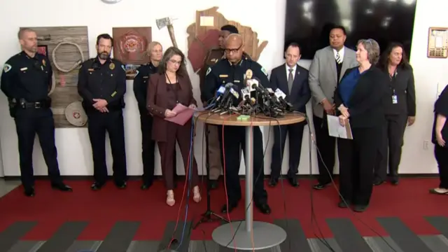 Shon Barnes speaking at a podium with microphones in front of him, four police officers and five other people in suits stand behind him