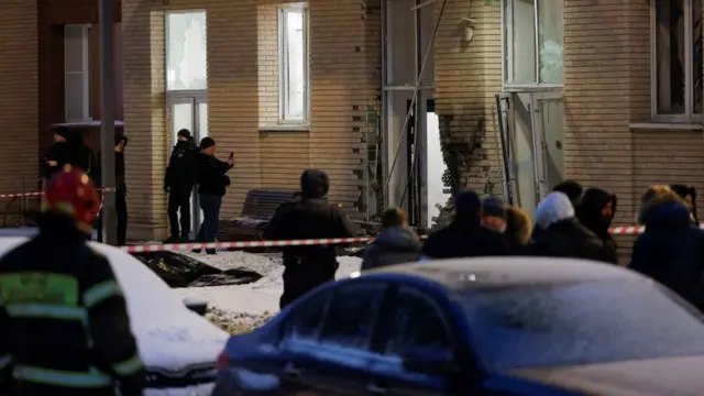 People stand behind a police cordon outside a damaged building. A black body bag can be partly seen on the snowy ground.