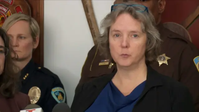 Satya Rhodes Conway speaking in front of microphones, two police officers in uniform are behind her