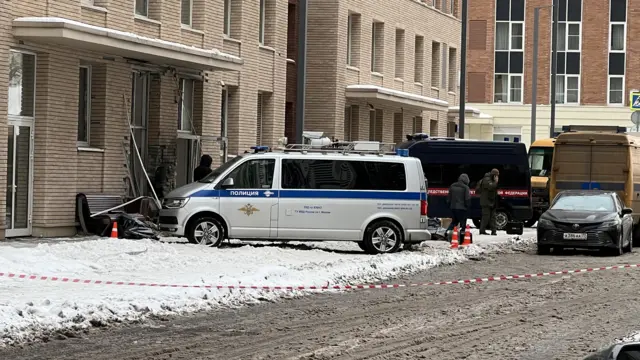 A police cordon around the apartment building. Behind the tape are emergency services vehicles and workers. Outside the damaged building is a bench and some debris. There is snow on the ground on the pavement and slush on the road.