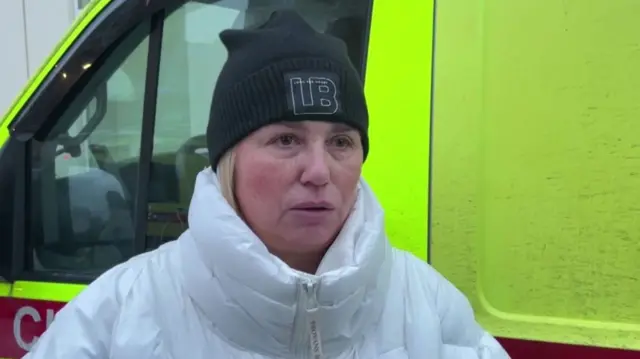 A woman in a white puffer coat and a black hat looks at the camera. An ambulance is behind her.