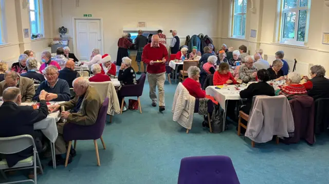 A group of people shares lunch together inside a church hall. The group is mostly made up of elderly men and women in winter clothing. A man in a red jumper and pink shirt walks in the centre of the room holding a plate, three men and a woman behind him queueing outside a kitchen to receive their food