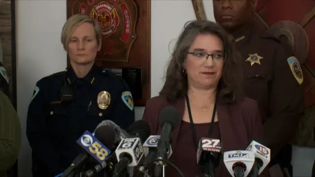 Melissa Agard speaking in front of many microphones, two police officers in uniform are standing behind them