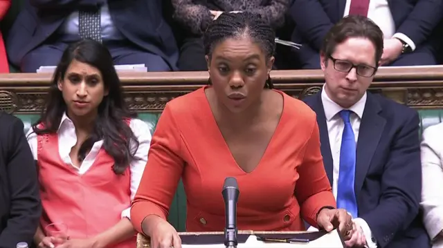 Kemi Badenoch stands at lectern asking questions during PMQs. She's wearing a red dress, her hands flipping sheets of paper. Sitting behind her, left from right, are Claire Coutinho MO and Alex Burghart