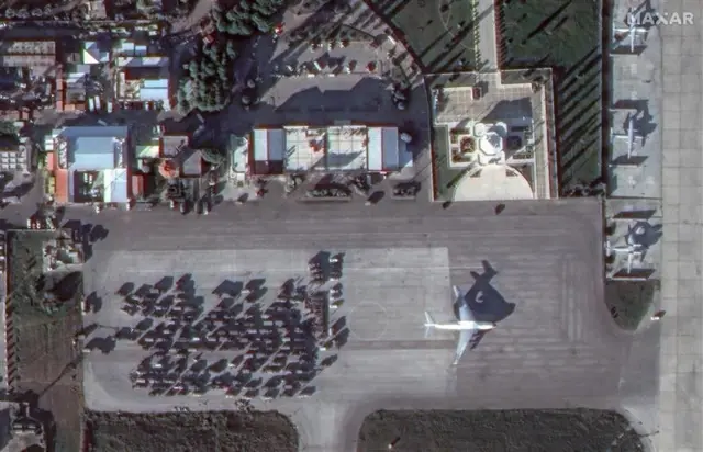 A satellite image of dozens of military vehicles parked in a Russian air base in Syria, a large aeroplane is visible - likely to air lift equipment out