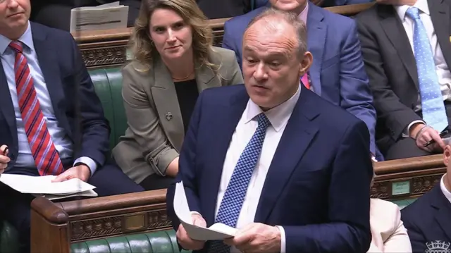 Ed Davey stands in House of Commons to ask questions during PMQs. He's wearing a dark blue suit, white shirt and patterned blue tie whilst holding a small stack of papers. Visible behind him are fellow MPs sitting on green leather benches