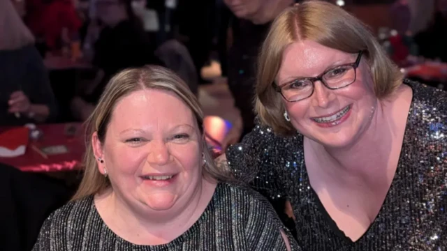 Laura and Alison smile wearing shiny and glittery black tops. On the right, Laura is wearing glasses with short hair. On the left, Alison smiles with blonde hair and small silver earrings.