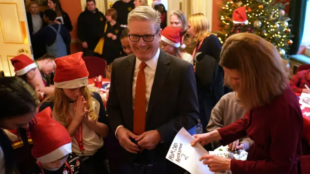 Starmer wears a suit and smiles down at a group of children wearing Santa hats while his wife Lady Victoria Starmer reads a piece of paper reading 'Mum, Dad Merry Xmas'