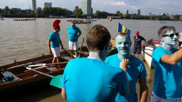 Men wearing blue t-shirts and painting themselves blue as the boats wait in the water