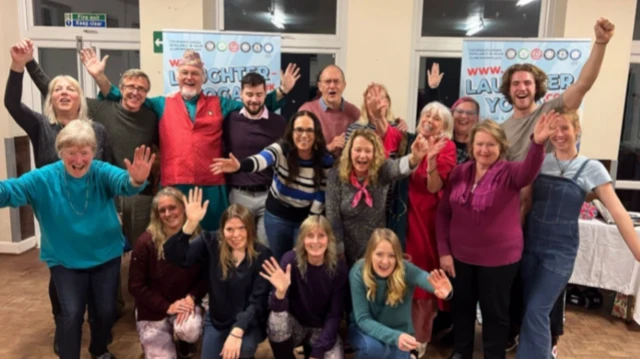 A group photo Clive St James' yoga class with around 18 people smiling with arms in the air