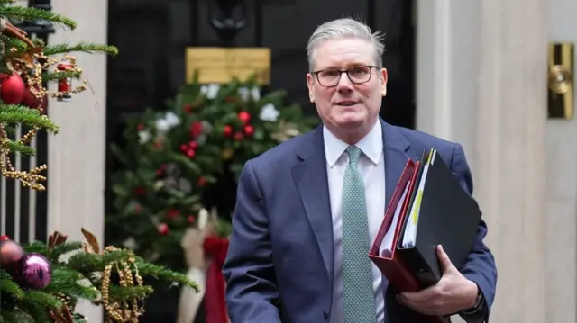 Keir Starmer leaves Number 10 to head to the Commons holding two folders under his left arm, one red and one black. Behind him is the black door of Number 10 decorated with a wreath. Branches of a Christmas tree are visible to Starmer's right