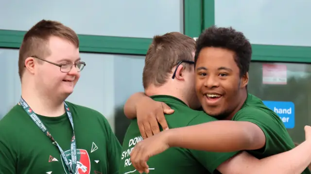 Three members of the Leicester Tigers rugby team for young people with Down's syndrome. Two members are hugging each other, with one smiling. Another member looks on with a smile. All are wearing green tops.