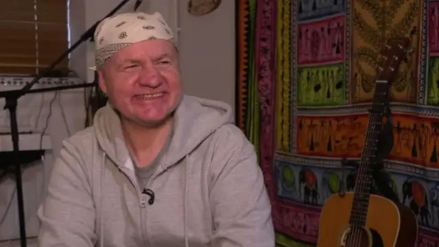 Shaun Hayward wearing a grey zip-up hoodie, smiling at the camera, with a guitar and microphone in the background