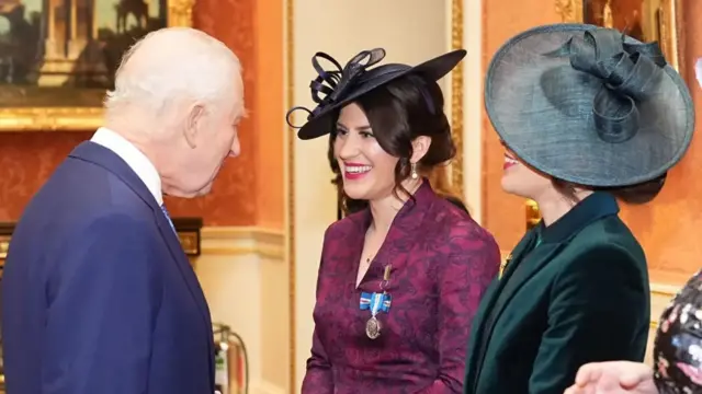 King Charles is talking with a woman with dark hair who is wearing a medal pinned to her purple dress and has a dark formal hat on her head. The medal has a blue, white and red ribbon. Alongside her is another woman who's face is obscured by a large hat. The photograph was taken inside Buckingham Palace.