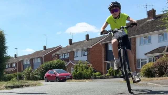 Dexter wearing a helmet and sunglasses rides on his bike on a residential road. He is wearing a yellow top and black shorts.