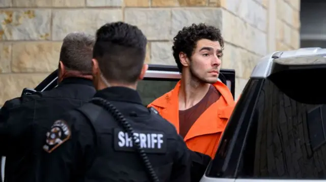 Luigi Mangione standing in an orange prison jumpsuit while two sheriff's face towards him as he leaves a car