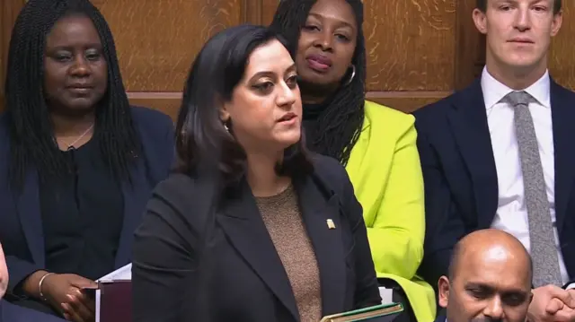 Labour MP Kirith Entwistle asks question to Keir Starmer from the benches during PMQs. She's in a black jacket and light brown jumper, colleagues are visible sitting behind her