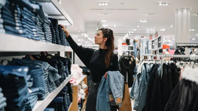 Young woman with brown hair in clothing store, looking at shelves stacked with jeans.