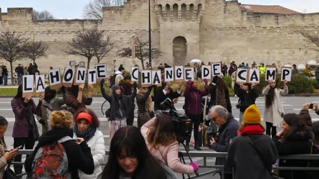 Women line up holding placards reading "la honte change de camp" ("shame changes sides")