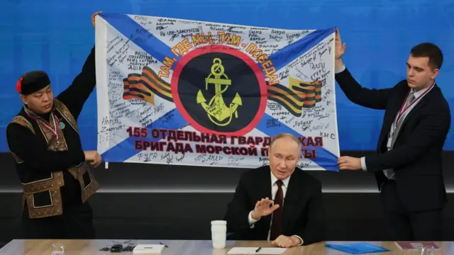 Putin sits at a table with a large military flag draped behind him.