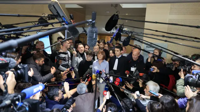 Gisèle Pelicot stands at the centre of a large crowd of journalists in the courthouse, with several microphones and cameras pointing at her