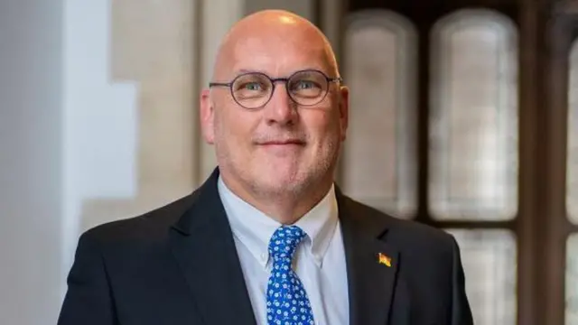 Nick Adams-King smiles at the camera. He is bald and wears a black jacket, white shirt, blue tie and round glasses