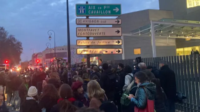 A crowd gathered beneath a road sign, beside a fence, in the early morning light