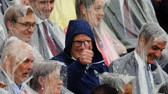 The prime minister, dressed in a branded navy blue raincoat, is surrounded by other spectators who are wearing throwaway waterproofs. He smiles at the camera and gives a thumbs up