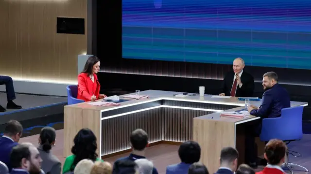 Three people sit around a horeshoe shaped desk during Vladimir Putin's end-of-year press conference. On the left, a woman with black hair in a bright-red suit looks down at  apiece of paper. Putin sits in the middle, one hand raised and on the right his spokesperson wears a blue suit looking out to the audiemnce. A blue screen stands behind them and there are rows of people sat in front.