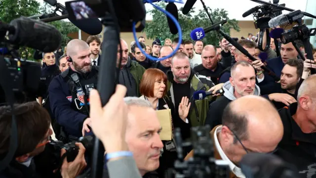 Gisèle Pelicot leaving the courthouse. She is surrounded by a large group of journalists, with mics and cameras pointed at her