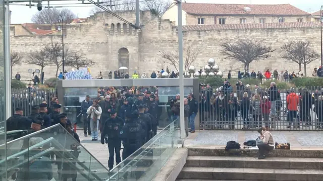 A crowd gathers at a gate and behind a fence at the courthouse. A number of police officers are standing within the courthouse grounds.