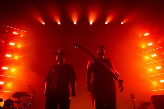 Femi and TJ Koleoso stood looking out at the crowd at Wembley Arena, with orange lights from the stage glowing around them