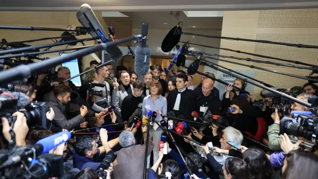 Gisèle Pelicot addresses a large crowd of journalists in the courthouse. She is flanked by her lawyers and family. there are several microphones being held up in front of her, including boom mics overhead