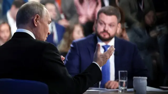 Putin gestures with his hand while a colleague sits across the table from him, in less focus.