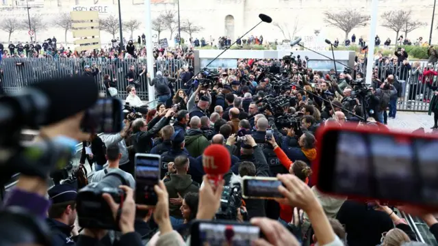 A large crowd fills the pathway that leads out of the courthouse to the street. Gisèle is walking somewhere in the crowd, only visible because of the boom mics overhead and the police surrounding her