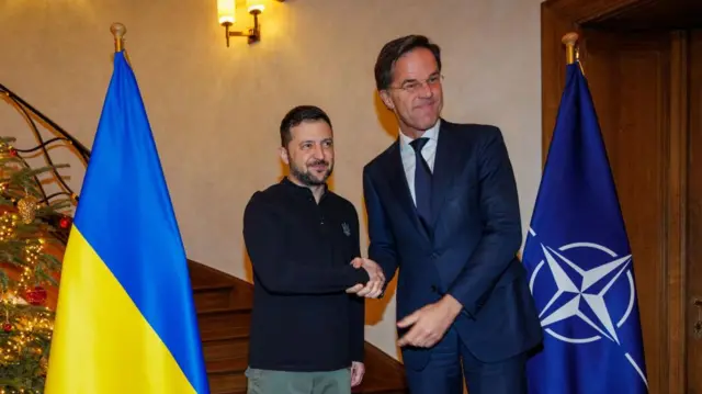 Ukrainian President Volodomyr Zelensky shaking hands with Nato cheif Mark Rutte. Rutte is on the right wearing a suit, Zelensky is wearing a blue jumper. There are Nato and Ukrainian flags wither side of them and a staircase behind them, a Christmas tree is partially visible.