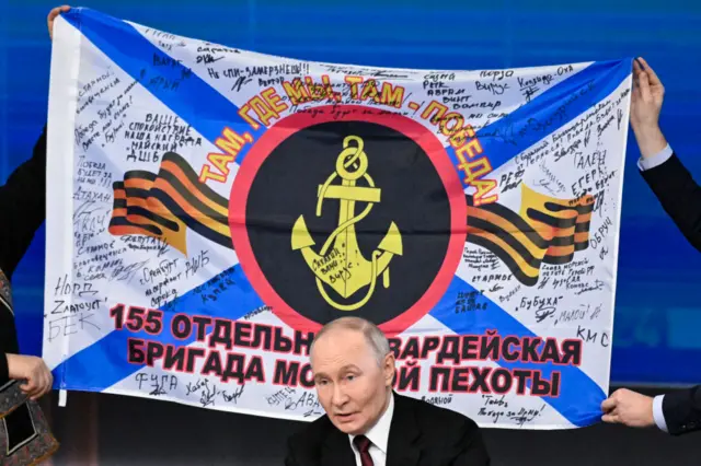 Putin sits at a table with a war banner behind his head