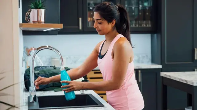 A woman wearing a pink top fills a water bottle from a tap