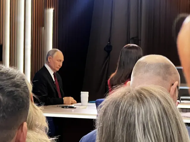 Vladimir Putin two rows of chairs away, sat at a desk