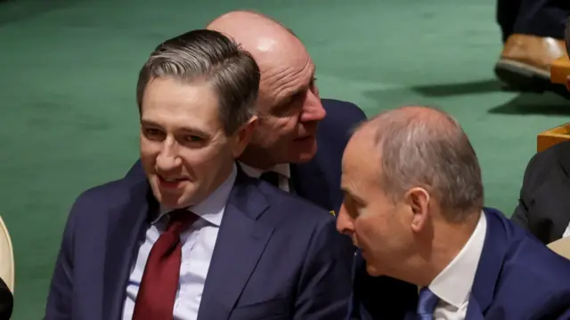 Simon Harris and Michael Martin sitting side-by-side, with Michael Martin leaning in to chat. Both are wearing blue suits