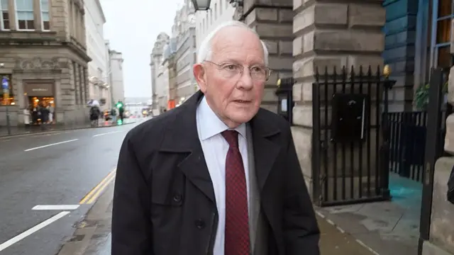 Sir Duncan Nichol walking along pavement after leaving the inquiry. It appears to be raining and he's wearing a dark grey suit, a maroon tie and wearing a thick black coat