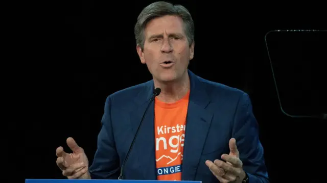 Greg Stanton, in an orange shirt and blue blazer talking into a microphone on a podium, with his hands raised