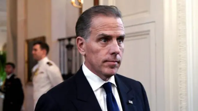 Hunter Biden, shot from chest up and wearing a dark suit and tie, inside the White House with three uniformed people blurred in background