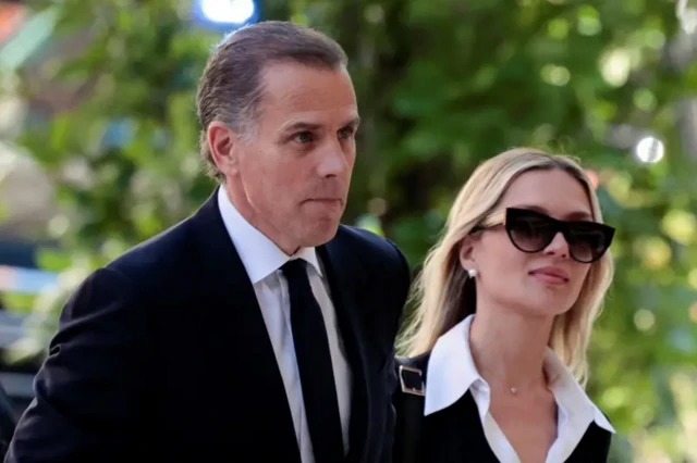 A photo taken form middle distance - Biden, a man with grey hair slicked back, walks past a tree wearing a suit. On his left, a blonde woman in sunglasses walks alongside him