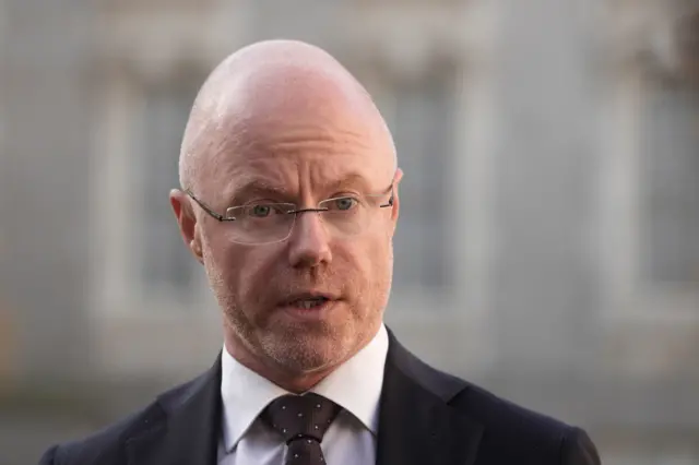 Stephen Donnelly is wearing a suit and tie. He is bald with some stubble and is wearing rimless glasses