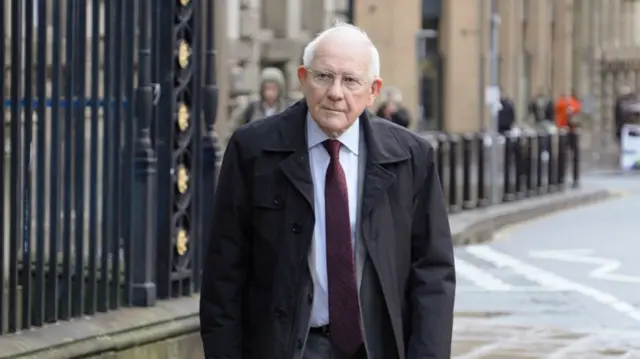 Sir Duncan Nichol walks along pavement to inquiry building. He's wearing a dark suit and a thick black coat.