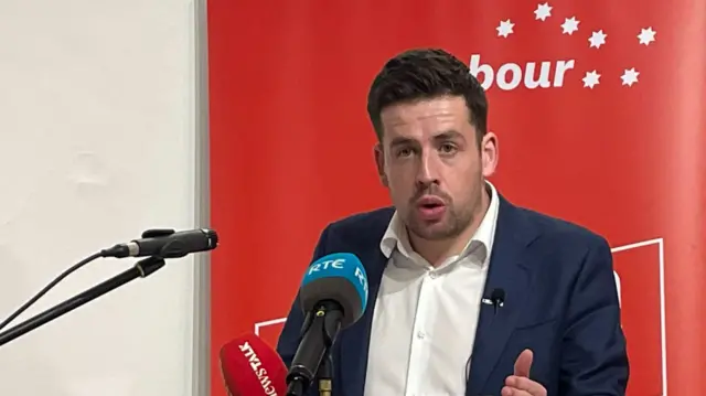 Labour's Conor Sheehan standing at a podium with microphones in front of him. He has dark hair and is wearing a blue suit.