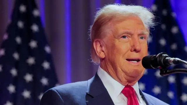 Donald Trump, on right side of image, shot from chest up, wearing a blue suit and red tie. Speaking into microphone with American flags blurred in background