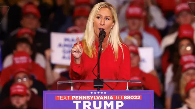 Taylor-Green, wearing a bright red jacket, points her finger as she speaks at a Trump campaign branded lectern to a rally crowd