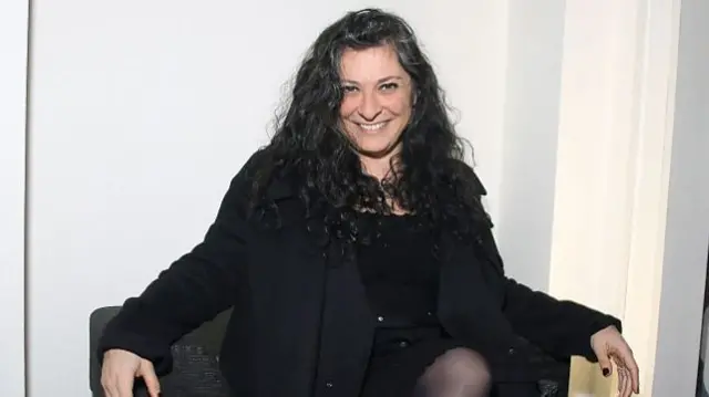 SOPHIA NEOPHITOU-APOSTOLOU sits in a chair. She has dark hair and wears a black outfit